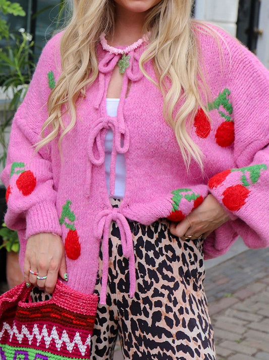 A woman wearing a vibrant pink knit cardigan adorned with red cherry appliqués and green leaf accents. The cardigan features an open-front design tied with delicate pink bows. 